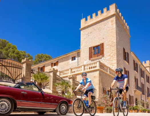 Two men biking and smiling, with a castle-like stone building in the background