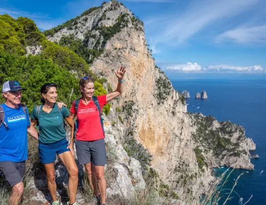 Two women and a man smiling and waving, while standing on a cliff next to the ocean