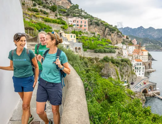 Two women and a man smiling while walking on a stone corridor on a hill, with the ocean to the right