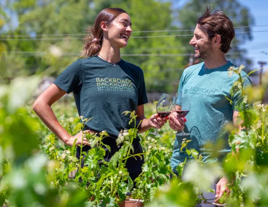 Man and woman smiling while holding up wine glasses, standing in a vineyard