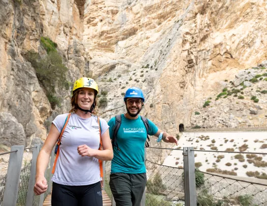 Man and woman walking on a wooden bridge with a large cliff in the background