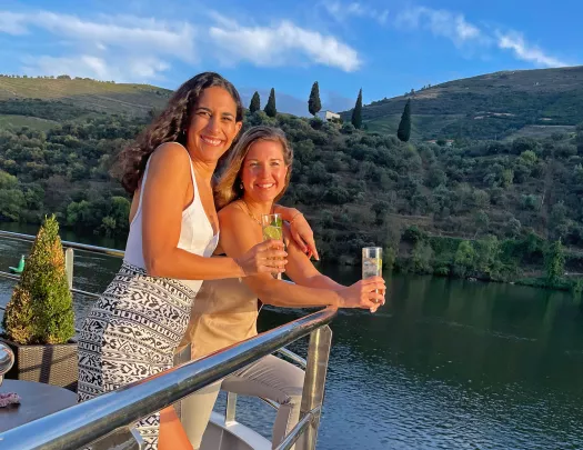 Two women smiling by a boat's railing, smiling while holding drinks
