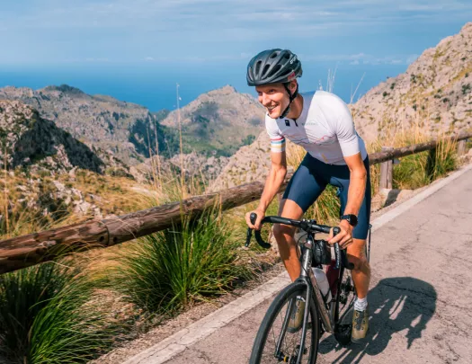 Man smiling while riding a bike on a road, with large cliffs in the distance