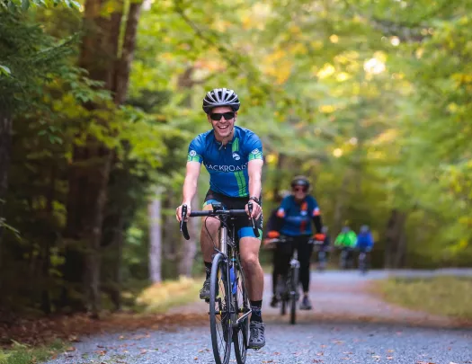 Man wearing sunglasses, riding a bike on a road surrounded by tall trees