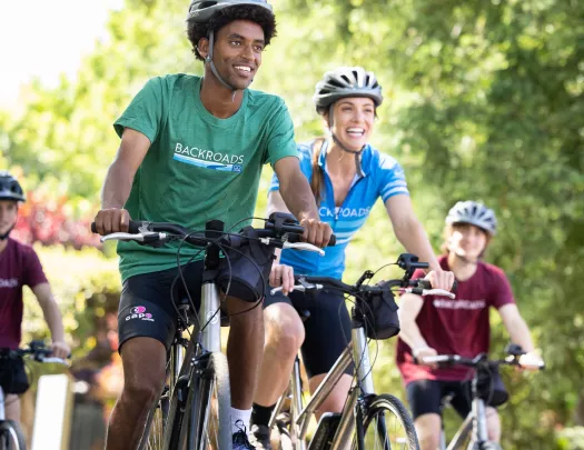 Group of men and women smiling while riding bikes on a road surrounded by tall trees