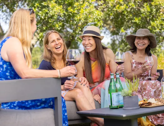 Four women outdoors, smiling and holding glasses of wine, with trees in the background