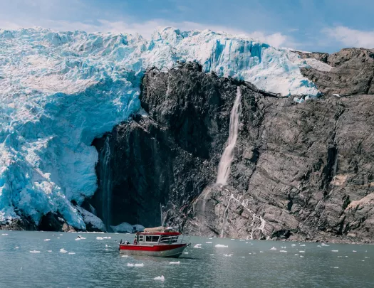 Red and gray boat floating next to large glaciers and cliffs