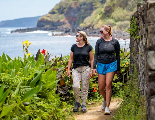 Two women smiling while walking on a dirt trail surrounded by large plants