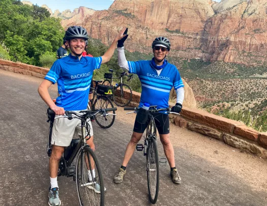 Two men wearing Backroads jerseys, high fiving each other and standing on their bikes
