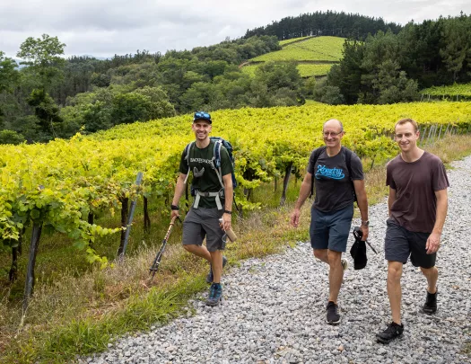 Three men smiling while hiking on a gravel trail, with crop fields in the background