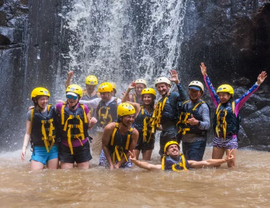 Group of people smiling while wearing helmets and life vests under a waterfall