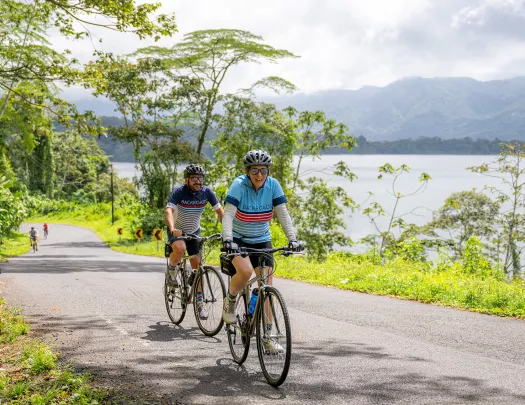 Two people riding bikes on a road, with a lake to the right