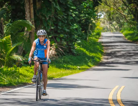 Woman wearing a blue and orange jersey, riding a bike on a road surrounded by tall plants