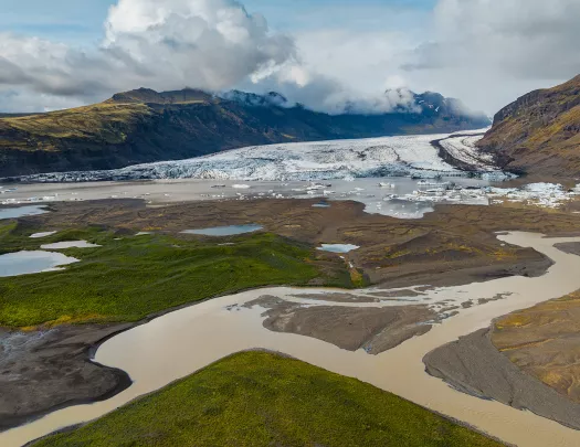 Valley with a brown river, in front of a large lake with ice caps behind it