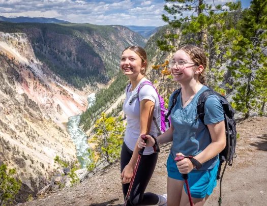Two girls using hiking poles, walking on a dirt trail and large mountains in the distance
