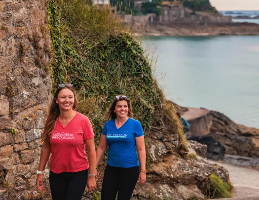 Two women walking along a stone back with hills and the ocean in the background