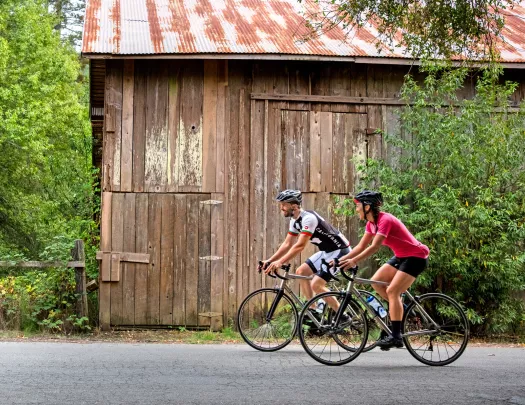 Man and woman wearing biking gear, riding bikes in front of a wooden barn on a road
