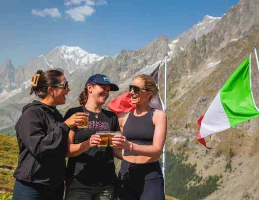 Three women smiling next to an Italian flag, with large mountains in the background