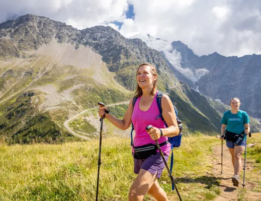 Two women walking on a dirt trail, with large mountains in the background