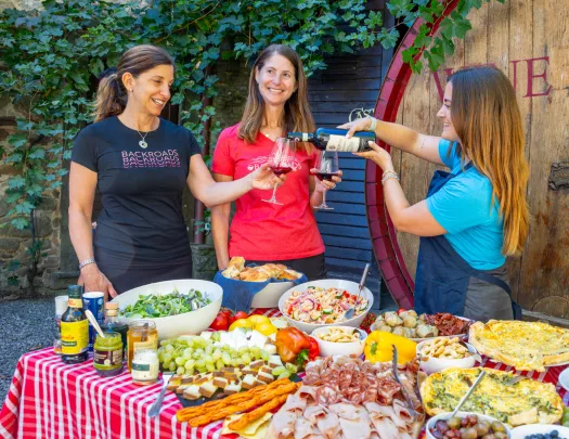 Three women behind a table full of food, pouring wine into glasses