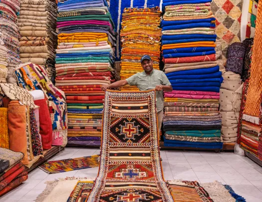 Man holding up a large rug in a room full of stacked rugs
