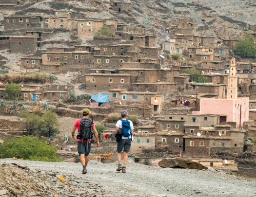 Two people walking on a dirt trail towards dirt and stone houses