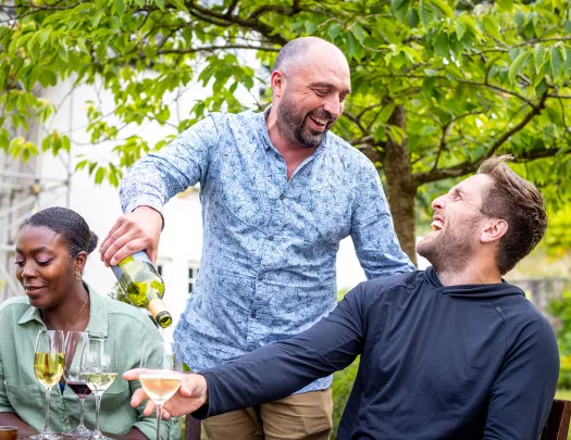 Man standing over a man sitting down, pouring him a glass of wine
