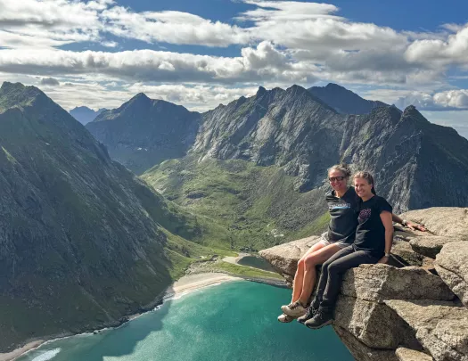 Two women sitting on the edge of a rock with a lake and mountains in the distance