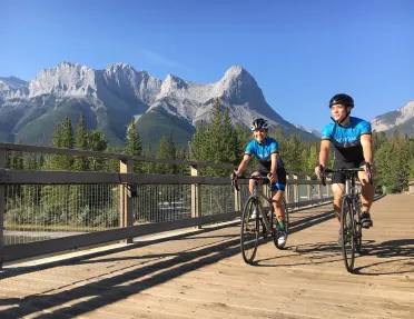 Two guests cycling on wooden path, Rockies in distance.