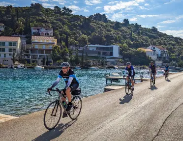 Four guests cycling down coastal road, blue ocean, hillside houses behind them.