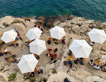 Overhead shot of people eating beneath white umbrellas, stone cliffs, ocean.