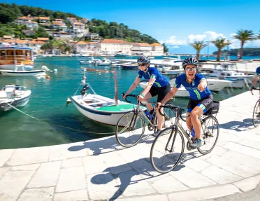 Three guests cycling on stone pier, boats, coastal town, ocean behind them.