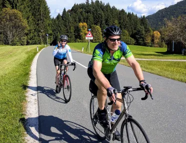 Two bikers riding down a hilly, tree lined street.