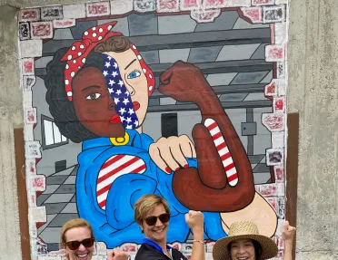 Three guests posing in front of Rosie the Riveter mural, doing her pose.