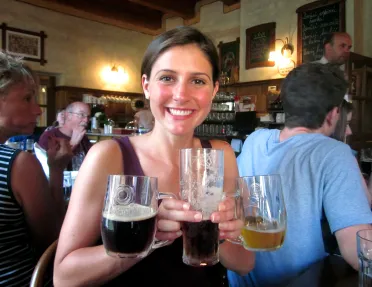 Woman posing in bar with three glasses of beer in varying colors.