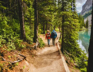 Two guests hiking down forest trail, river or lake to their right.