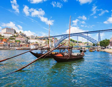 Two dark wood boats on Douro River, large metal bridge, colorful houses behind.