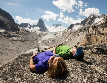Two guests laying on rocky mountainside, arid cliffs and clouds in background.