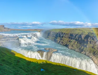 Wide shot of alpine vista, waterfall, rainbow, cliffs on either side. 