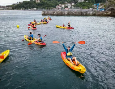 Group of guests in kayaks, one raising paddle over head, coast behind them.