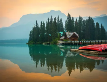 Wide shot of large lake, wooden house, trees, canoes, mountains visible.