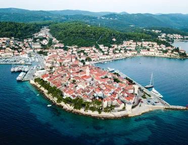 Wide shot of coastal Croatian town, white and tan buildings, boats, forest in distance. 