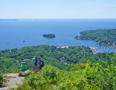 Wide shot of a coastal vista, sailboats visible in water, group of young guests in foreground.