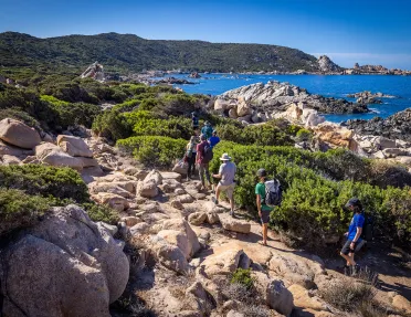 Eight guests hiking on rocky shore, ocean to their right, hills in distance.