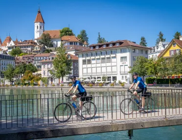 Two guests cycling over concrete bridge, Thun cityscape in distance.