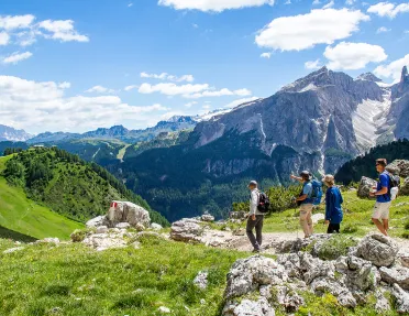 Group of guests hiking down mountain trail, Dolomite range in background.