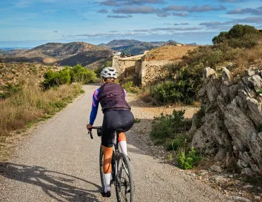 Guest cycling down gravel road, towards stone ruins, golden hills. 