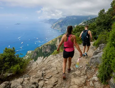 Two guests hiking down cliffside, boats in ocean down below.