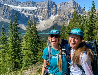 Two guests smiling for camera, snowy mountain, trees, blue sky in background.