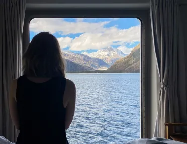 Guest on cruise ship in room, looking out towards mountains.